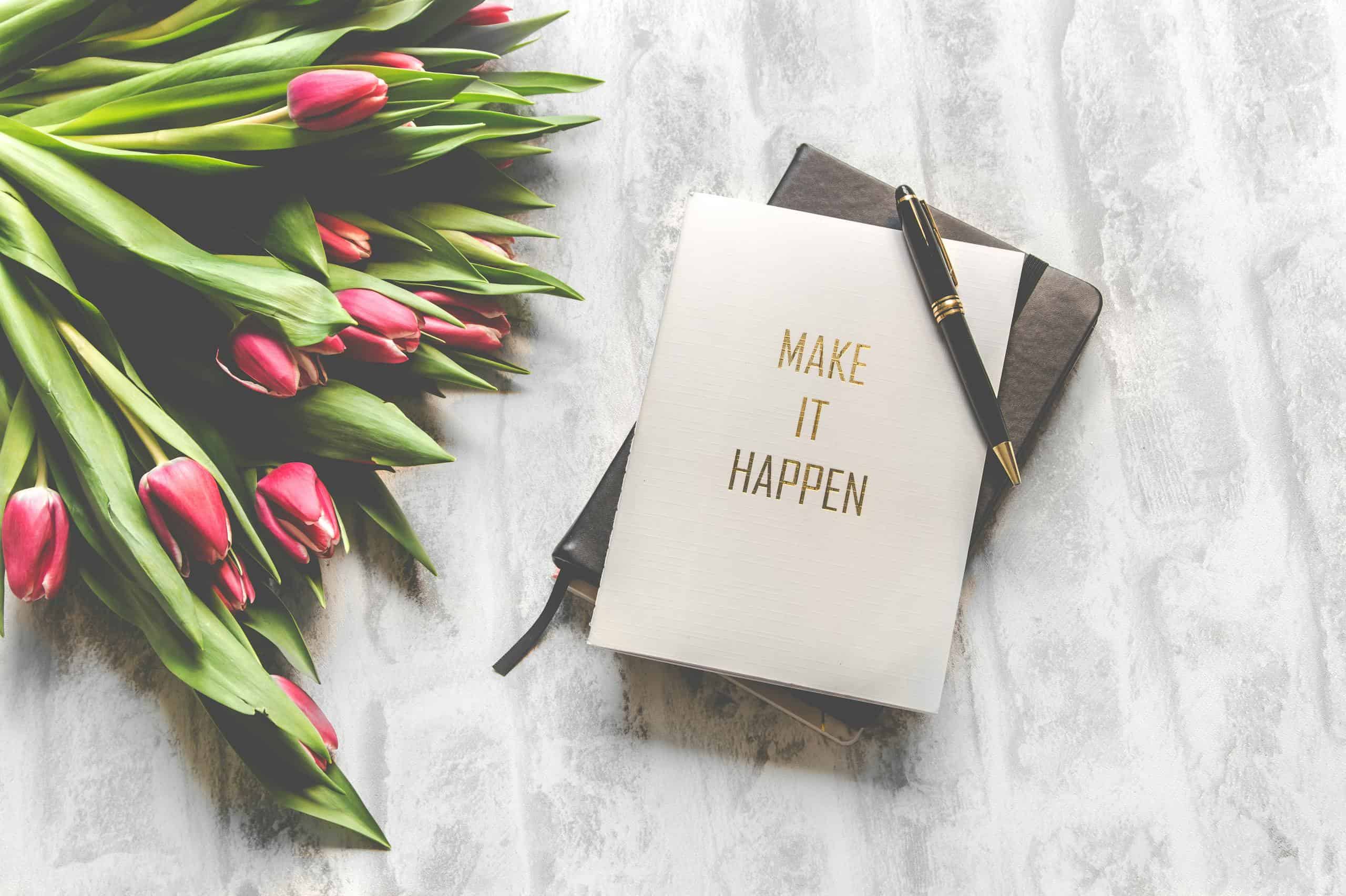 A notebook with 'Make It Happen' inscription beside a bouquet of pink tulips, capturing inspiration.